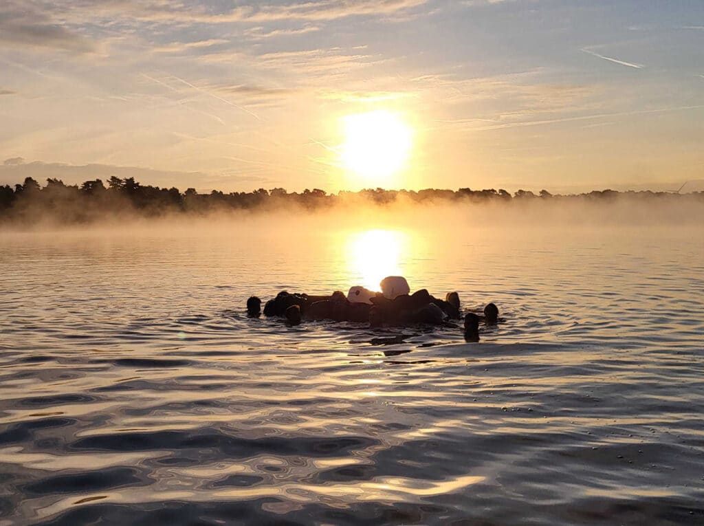 Een serene zonsopgang boven een meer, waarbij een groep mensen in een cirkel op het water drijft. De mist hangt laag boven het water, wat een rustige en mysterieuze sfeer creëert.