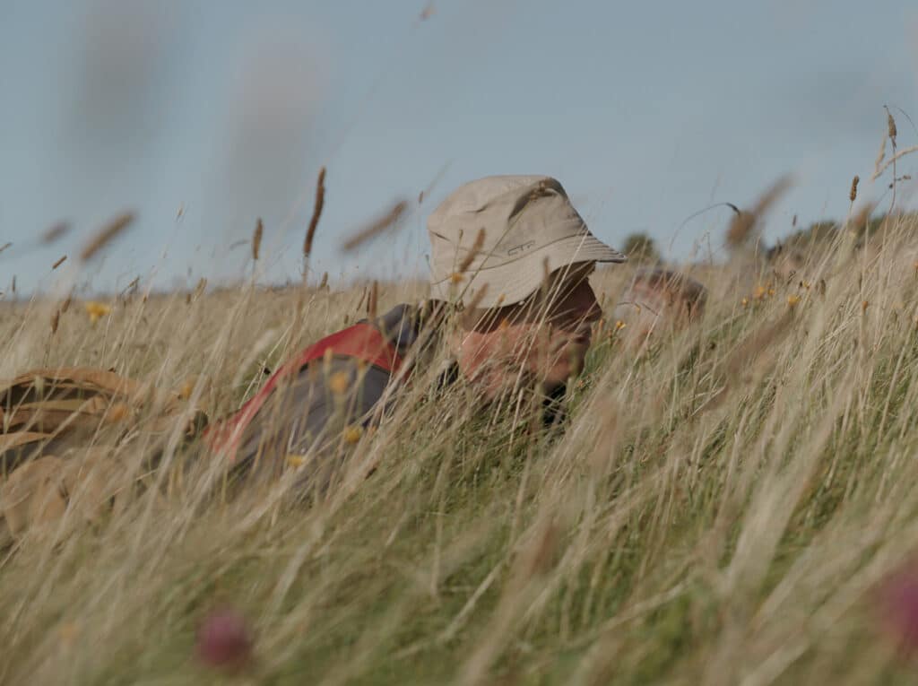 Een persoon in een hoed kruipt door hoog gras, waarschijnlijk tijdens een militaire oefening. Het gras en de wilde bloemen zorgen voor een natuurlijk camouflagescherm.